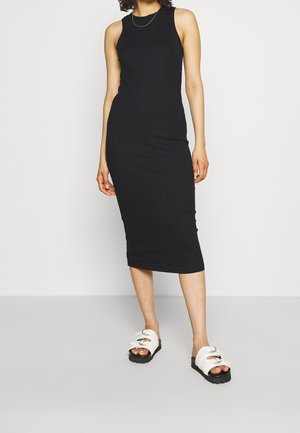 Kvinde iført en ærmeløs, knælængde sort kjole og hvide platformsandaler med dobbelte spænder, stående foran en ensfarvet lys baggrund.