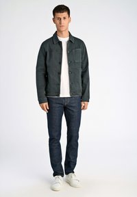 Chaqueta de mezclilla oscura con bolsillos frontales y cierres de botón, llevada sobre una camiseta blanca. Combinada con jeans oscuros y zapatillas blancas.