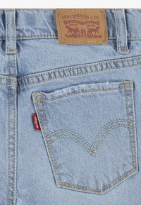 Světle modré džíny vyrobené z denimu s klasickým designem zadní kapsy, žlutým prošíváním a hnědou koženou etiketou s červeným detailním štítkem.