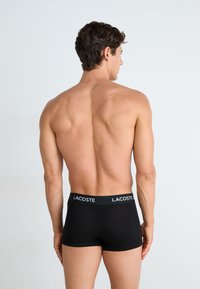 Schwarze Boxershorts für Herren aus weichem Material mit einem breiten Bund, der das weiße "LACOSTE"-Logo trägt. Von hinten betrachtet.