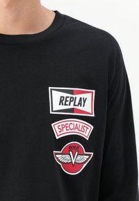 Black shirt with three red, white, and black patches reading "REPLAY," "SPECIALIST," and a winged "V" logo on the chest.