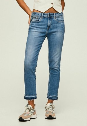 Frau trägt blaue, mittelhohe kurze Jeans mit ausgefransten Säumen und beigen Sneakers, gezeigt vom Bauch bis zu den Füßen vor einfachem Hintergrund.