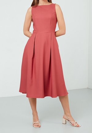 Vrouw draagt een mouwloze, nauwsluitende, koraalkleurige midijurk met geplooide rok, gecombineerd met witte sandalen met hak, staand tegen een effen achtergrond.