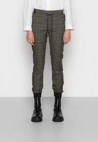 Person klädd i grå rutiga joggarbyxor med svart dragsko, svarta ankelkängor och en vit långärmad tröja som står mot en enfärgad bakgrund.