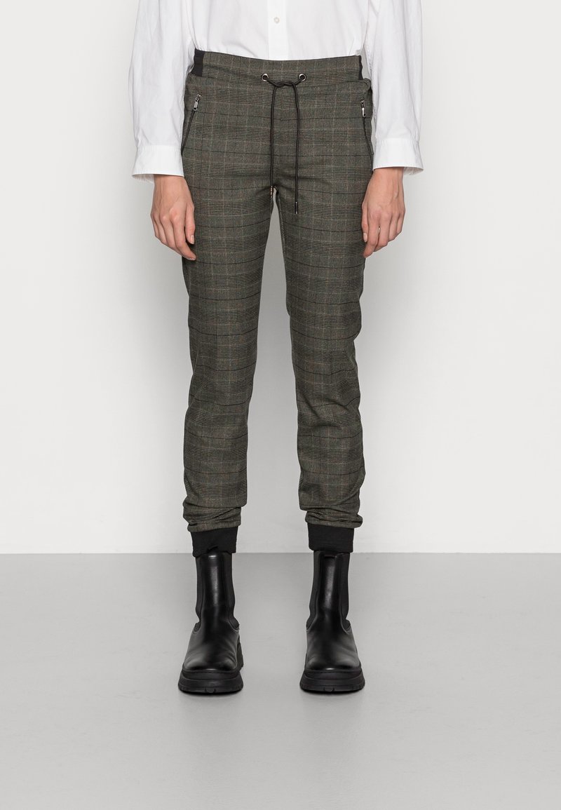 Person klädd i grå rutiga joggarbyxor med svart dragsko, svarta ankelkängor och en vit långärmad tröja som står mot en enfärgad bakgrund.