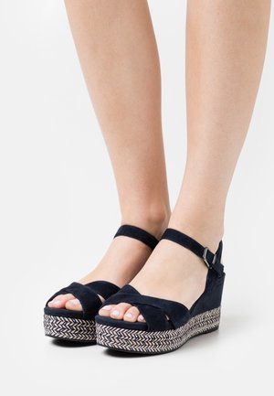 Sandales compensées en suede marine avec des brides croisées, une sangle de cheville avec boucle et une plateforme en chevron tissée. Design à bout ouvert.