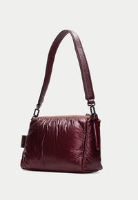 Sac à main bordeaux avec une surface lisse et texturée. Possède une longue bandoulière en cuir réglable et une fermeture éclair en haut.