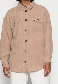 Beiges Cordhemd mit durchgehender Knopfleiste, zwei Brusttaschen und spitzem Kragen. Mit schwarzen Knöpfen und langen Ärmeln.
