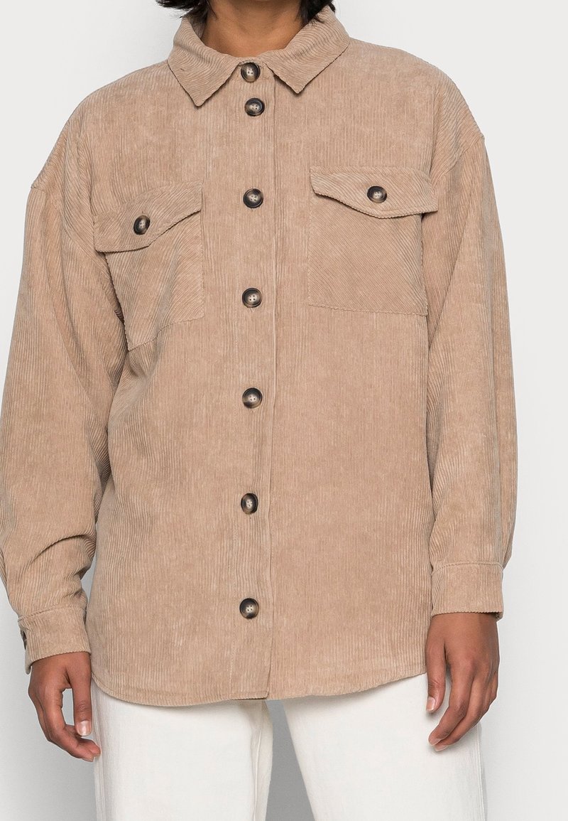 Beiges Cordhemd mit durchgehender Knopfleiste, zwei Brusttaschen und spitzem Kragen. Mit schwarzen Knöpfen und langen Ärmeln.