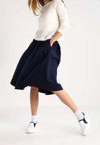 Marineblauwe midi-rok met zakken, gecombineerd met een witte gebreide trui en witte instap-schoenen met zwarte accenten, rustend boven witte sokken.