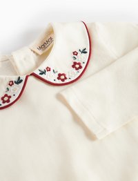 Cremfarbendes Langarmshirt mit einem rot eingefaßten Kragen, der mit floralen Stickereien in Rot, Weiß und Grün verziert ist. Das Etikett ist innen sichtbar.