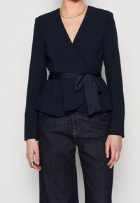 Námořnická blůza s dlouhými rukávy, peplum designem a zavázaným páskem. Hladká tkanina s hlubokým V výstřihem. Nosená s tmavými džíny.