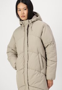 Casaco puffer bege com capuz, apresentando seções almofadadas, bolsos laterais e um fecho éclair frontal. Feito de um tecido macio e resistente à água.