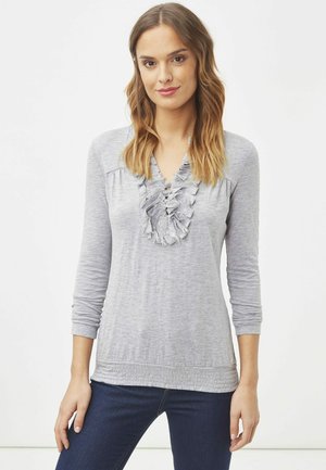 Mujer con cabello castaño claro que lleva una camiseta de manga larga gris claro con cuello con volantes y jeans oscuros, de pie frente a un fondo blanco liso.
