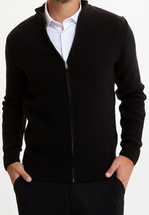 Mann trägt eine schwarze, strukturierte Strickjacke mit Reißverschluss über einem weißen Hemd mit Knöpfen, Hände in den Taschen einer schwarzen Hose, vor schlichtem Hintergrund.