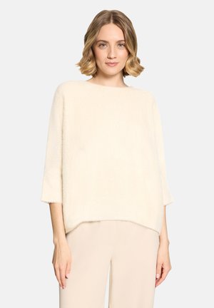Kvinde med kort blondt hår iført en løs cremefarvet, plysset sweater og lyse beige bukser, stående mod en ensfarvet baggrund.