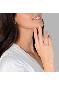 Anello d'argento con una pietra preziosa rotonda. La fascia è liscia e lucidata, posizionata su una mano che tocca il viso. Pelle chiara e tessuto bianco visibili.