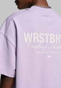 Levanduľové oversized tričko s bielym grafickým textom na zadnej strane, ktoré obsahuje slová "WRSTBH" a "Crafting Aesthetic", a má hladkú bavlnenú štruktúru.