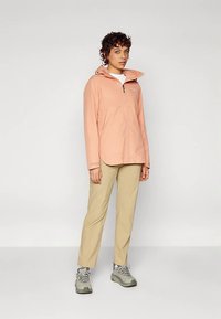Donna in piedi con una giacca zip-up pesca, pantaloni beige, scarpe da trekking grigie e una camicia bianca su uno sfondo neutro.