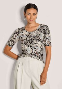 Vrouw in een zwart bloemenpatroon kortekorte top, ingestopt in hoge witte plooibroek, poseert met één hand op haar heup.