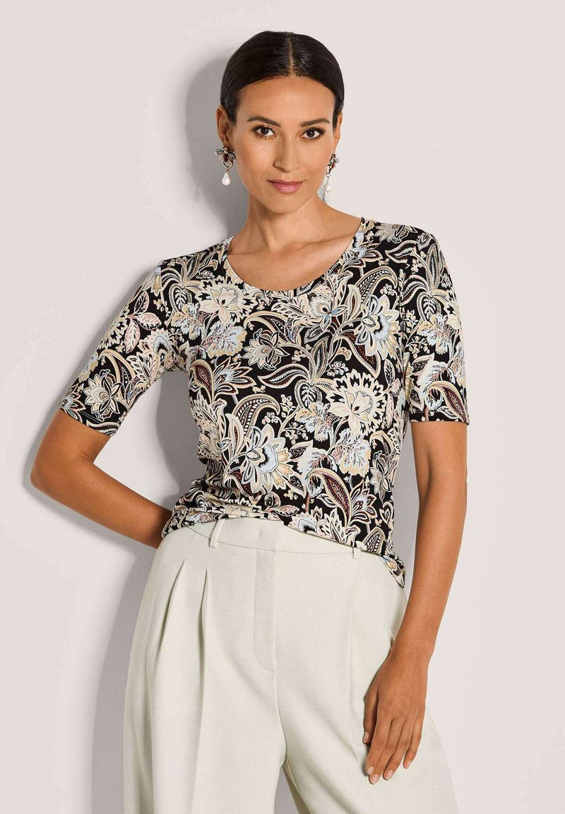 Vrouw in een zwart bloemenpatroon kortekorte top, ingestopt in hoge witte plooibroek, poseert met één hand op haar heup.