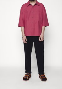 Košile s krátkým rukávem na knoflíky v jednobarevné bordó barvě, kombinovaná s tmavými kalhotami. Obuv má černý základ s barevnými pásky a doplňky.