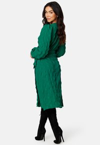 Grünes, strukturiertes Kleid mit langen Ärmeln, einer taillierten Taille und einem knielangen Saum. Kombiniert mit schwarzen, wildledernen Stiefeln über das Knie.