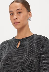 Top nero con texture e un effetto metallizzato, caratterizzato da un colletto a chiave e maniche lunghe. Abbinato a orecchini scultorei argentati.