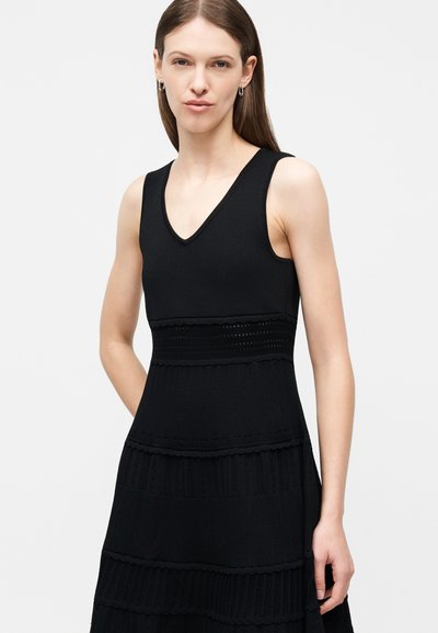 Femme aux longs cheveux bruns portant une robe noire sans manches à encolure en V avec des bandes horizontales texturées, debout devant un fond gris clair.