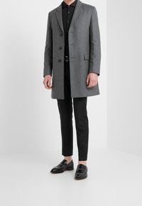 Homme portant un manteau gris mi-genou par-dessus une chemise noire, un pantalon noir slim et des mocassins en cuir noirs, debout devant un fond blanc.