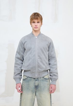 Persona joven con cabello rubio corto lleva una chaqueta gris claro con cremallera y vaqueros azules desgastados, de pie sobre un fondo blanco liso.