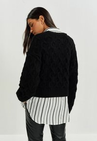 Frau mit langen dunklen Haaren, die einen schwarzen strukturierten Pullover über einem weiß-schwarz gestreiften Hemd und schwarzen Lederhosen trägt, und sich leicht abwendet.