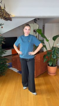 Μπλε κοντομάνικο μπλουζάκι με λογότυπο, μαύρο παντελόνι φαρδιάς γραμμής και ασημένια παπούτσια, στέκεται σε ξύλινο πάτωμα με φυτά και έπιπλα στο φόντο.