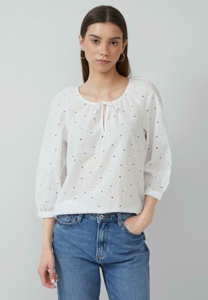 Weiße Bluse mit rundem Ausschnitt, Knoten-Detail und floralem Lochmuster. Lange, geraffte Ärmel und lockere Passform. Getragen mit blauen Jeans.