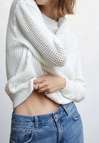 Biały, dziany sweter o luźnym kroju i otwartej strukturze, w połączeniu z niebieskimi dżinsami o wysokim stanie. Na jednej ręce znajduje się niebieski pierścionek.