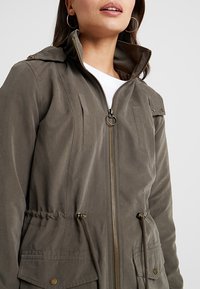 Khaki jas met capuchon, ritssluiting aan de voorkant en trekkoord in de taille. Voorzien van twee borstzakken en knoopaccenten. Zachte, gestructureerde stof.