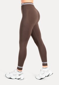 Leggings marron taille haute avec une texture lisse et une bande blanche contrastante en haut, associés à des chaussures de sport blanches et noires.
