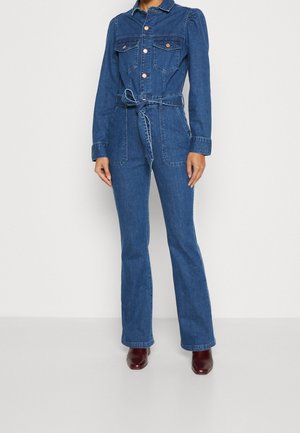 Femme portant une combinaison en denim bleu avec des poches avant, des poches poitrine à boutons, une ceinture nouée à la taille et des bottines marron.