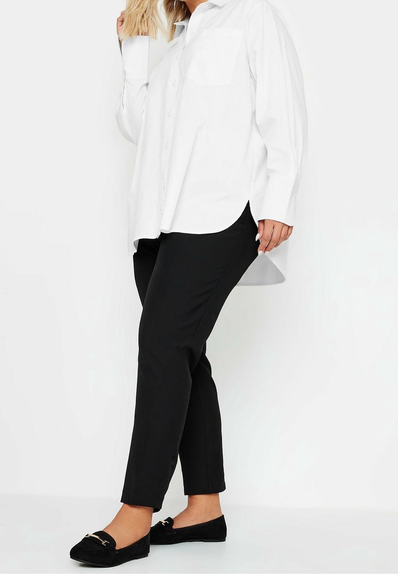 Woman wearing a loose white button-up shirt, black slim pants, and black loafers standing against a plain white background.