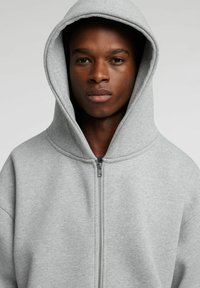 Grå hettegenser med glidelås laget av mykt stoff, med stor hette og avslappet passform med synlige sømmer. Enkel design uten mønstre.