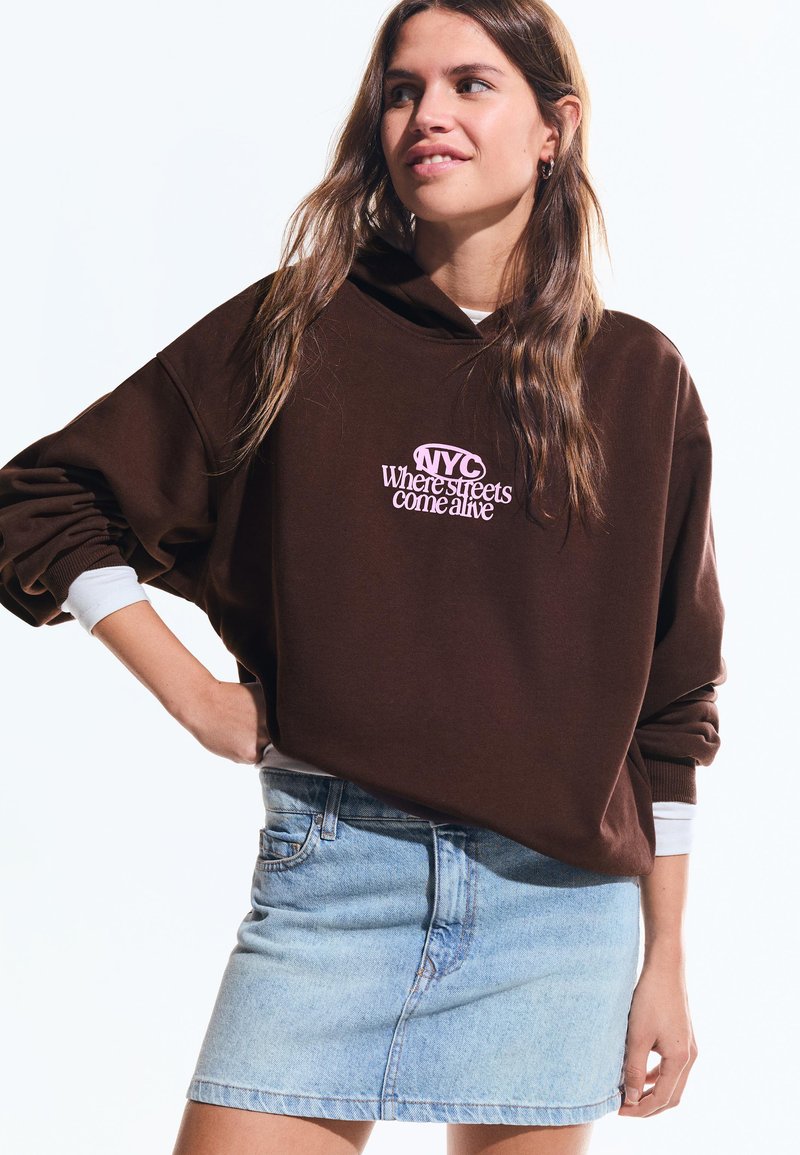 Jonge vrouw in een bruine hoodie met de tekst "NYC Where streets come alive", draag een lichte denimrok, glimlachend en naar de zijkant kijkend.