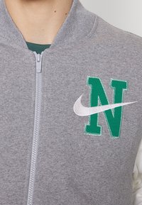 Felpa grigia con zip, dotata di un'emblema verde "N" e logo Nike bianco. Presenta un colletto a coste e un tessuto testurizzato.