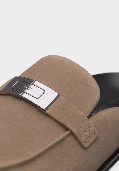 Μπεζ μοκασίνι από σουέτ με λεπτομέρεια ασημένιας μεταλλικής αγκράφας και μαύρη εσωτερική επένδυση, παρουσιάζοντας κοντινή λήψη του ανώτερου μέρους και του ιμάντα του παπουτσιού.