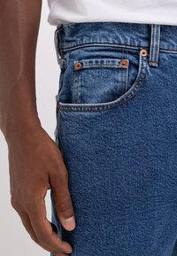 Vaqueros de mezclilla azul con una vista en primer plano de la zona del bolsillo, que presenta remaches de cobre, costuras en contraste y una textura suave.