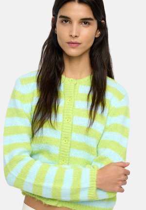 Kobieta o długich ciemnych włosach, ubrana w miękki, zapinany na guziki kardigan w poziome paski w pastelowej zieleni i jasnym błękicie.