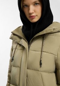 Khaki pufferjack die een hoge kraag, een ritssluiting aan de voorkant en trekkoorden heeft. Gecombineerd met een zwarte gebreide capuchon voor extra warmte.