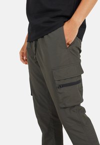Zielone oliwkowe spodnie cargo wykonane z lekkiego materiału. Charakteryzują się wieloma kieszeniami, elastycznym pasem ze sznurkiem oraz detalami na zamek.