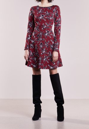 Femme portant une robe rouge à fleurs à manches longues associée à des bottes noires à talons montant jusqu'au genou, debout devant un fond uni.