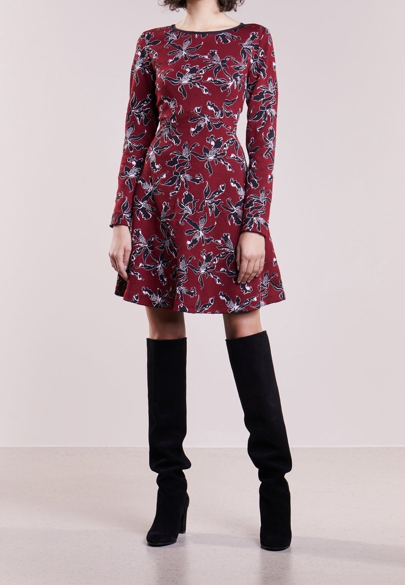 Femme portant une robe rouge à fleurs à manches longues associée à des bottes noires à talons montant jusqu'au genou, debout devant un fond uni.