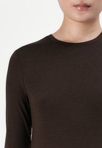 Persoană îmbrăcată cu un tricou simplu, de culoare maro închis, cu mâneci lungi, văzută de la gât până la jumătatea toracelui, pe un fundal alb.